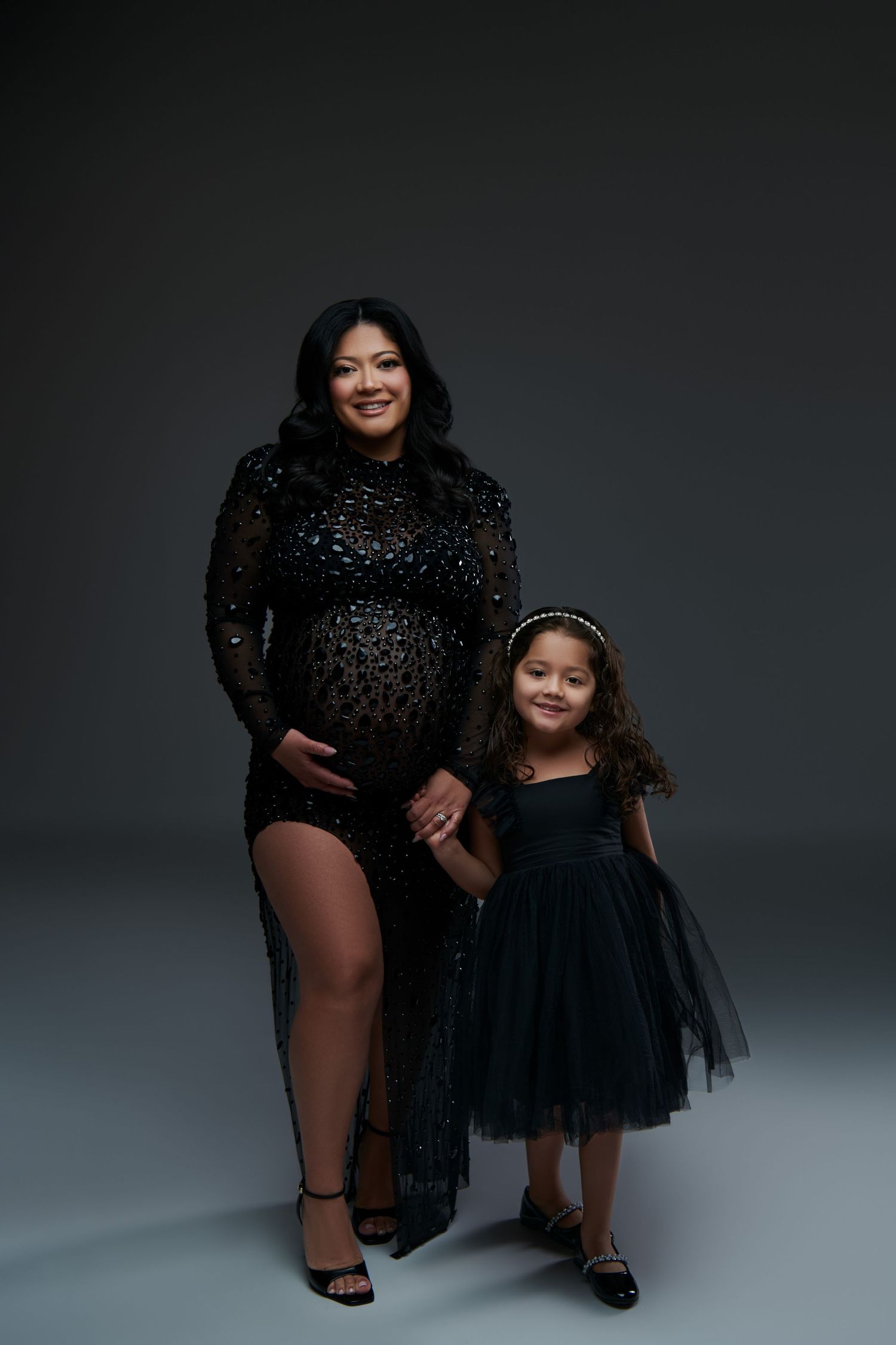 Maternity portrait showing woman in sparkly black dress with young girl in matching tulle dress against dark backdrop. NJ Luxury Maternity