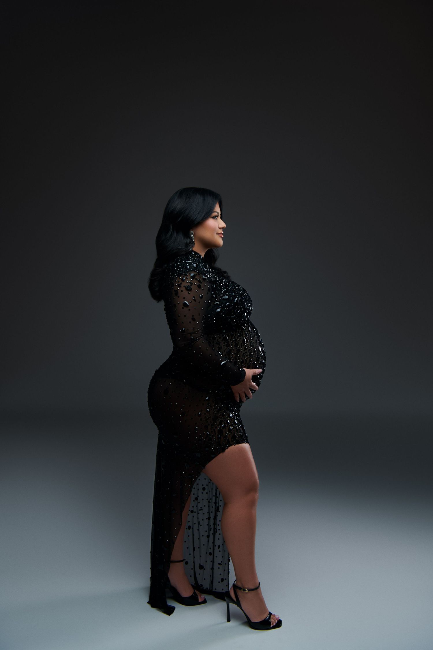 A dramatic portrait in a sheer black sequined gown with side slit against dark background. Gricel's Photography Maternity Photographer