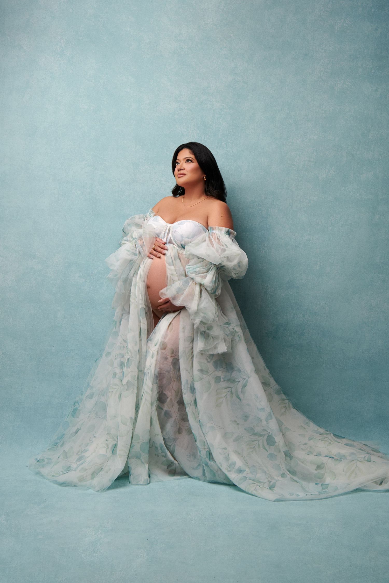 A flowing sheer white maternity gown flows gracefully against a muted blue studio backdrop. NJ Luxury Maternity Photographer