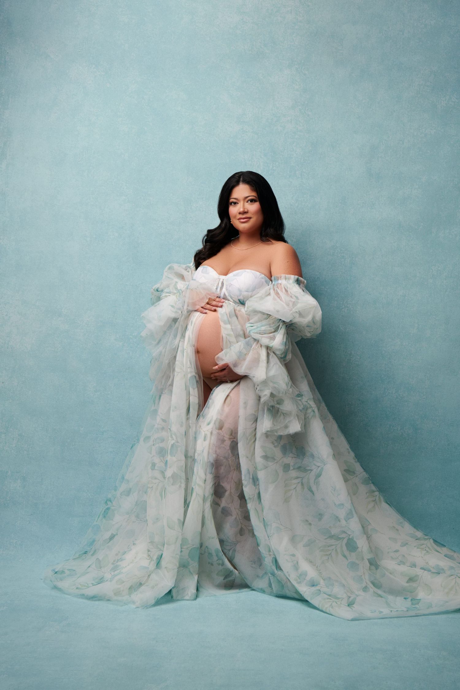 An ethereal maternity portrait features a billowing white gown against a soft blue background. Gricel's Photography Luxury Maternity Photos