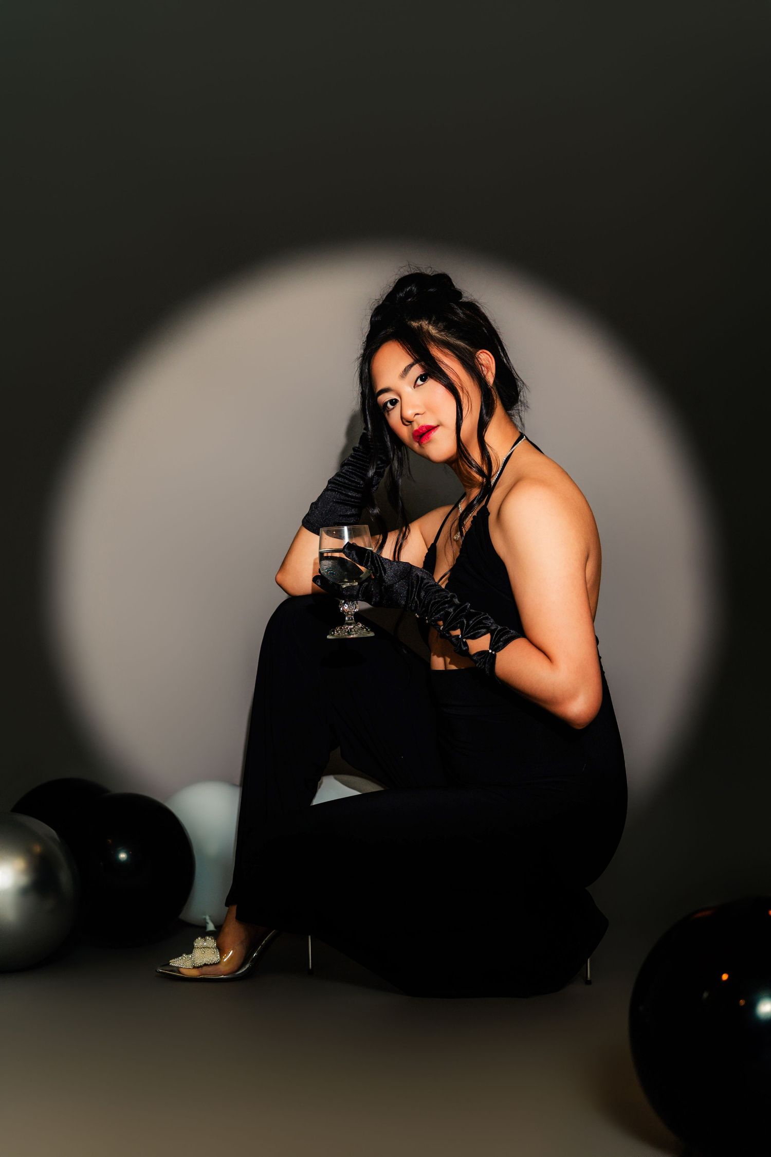 A figure in a black dress and sunglasses poses dramatically against a dark background with oval lighting effects.