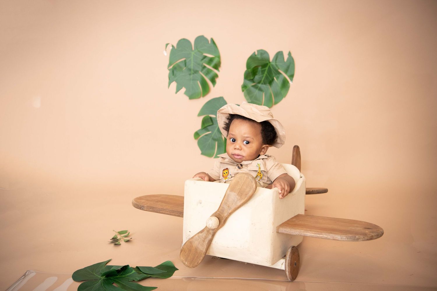 A toddler sits in a wooden toy airplane with green monstera leaves scattered against a beige background.