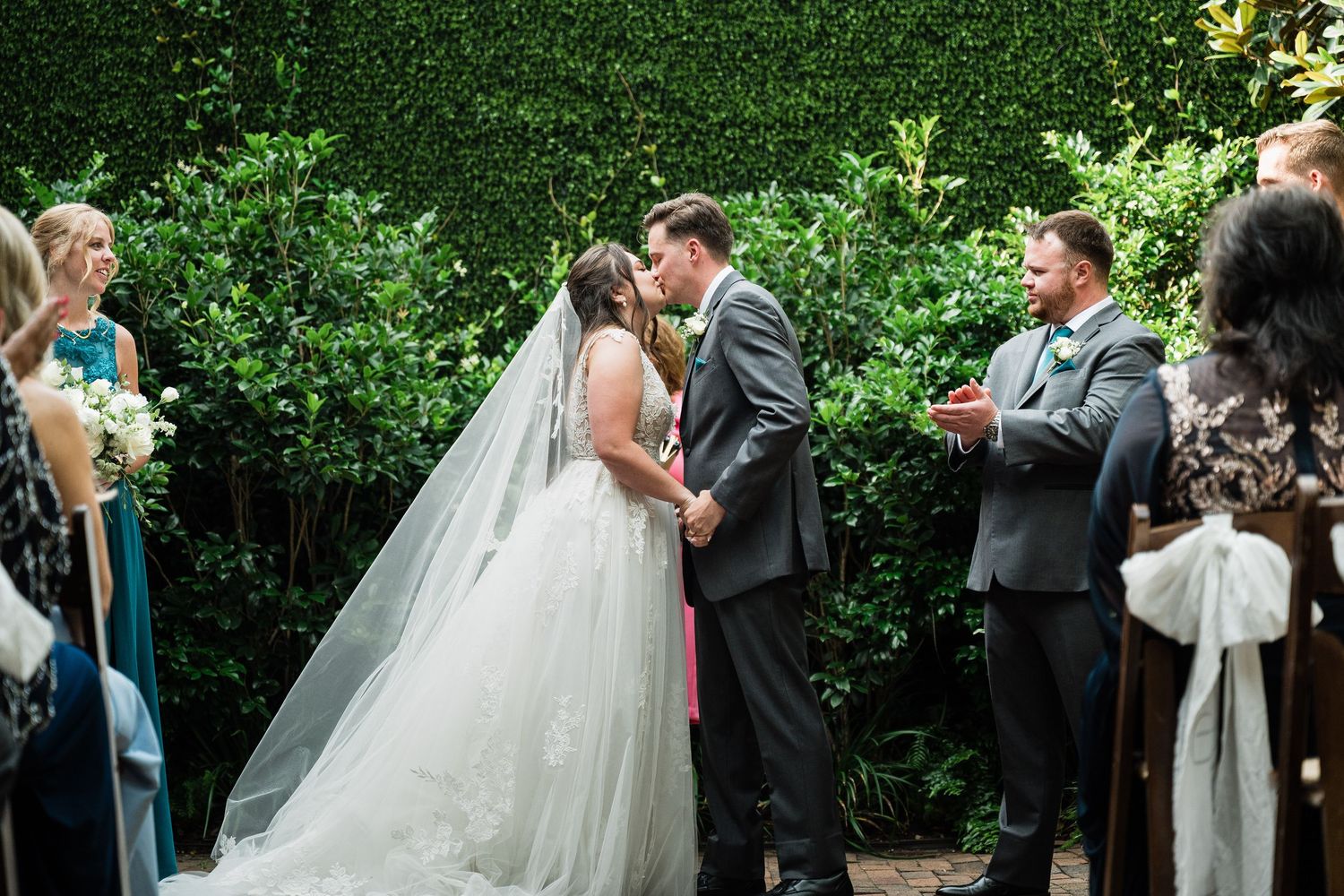 Couple shares romantic first kiss during outdoor garden wedding ceremony as guests look on.