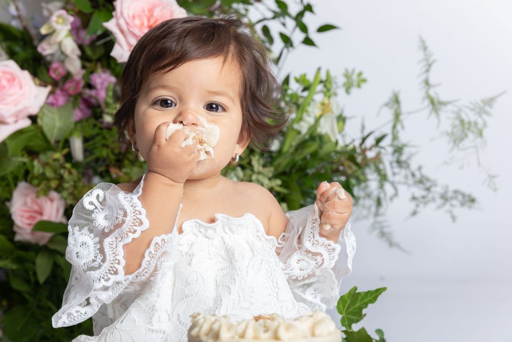 Baby in white lace dress and bow sits amid pink roses and greenery for a professional portrait session.