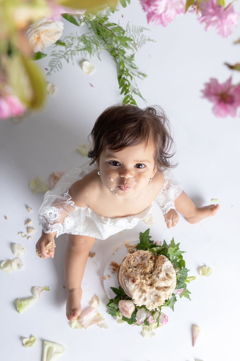 Overhead view of cake smash photography session with pink flowers and frosting scattered around white backdrop.