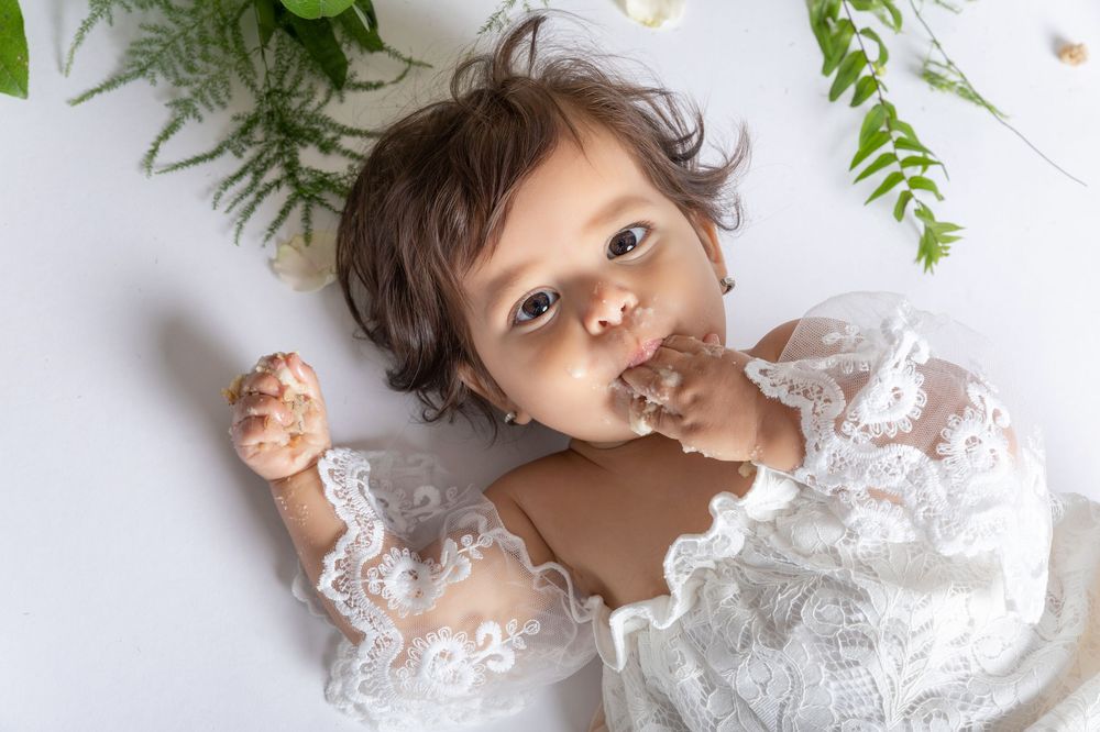 A baby wearing white lace lies on a white background with green foliage decorations overhead.