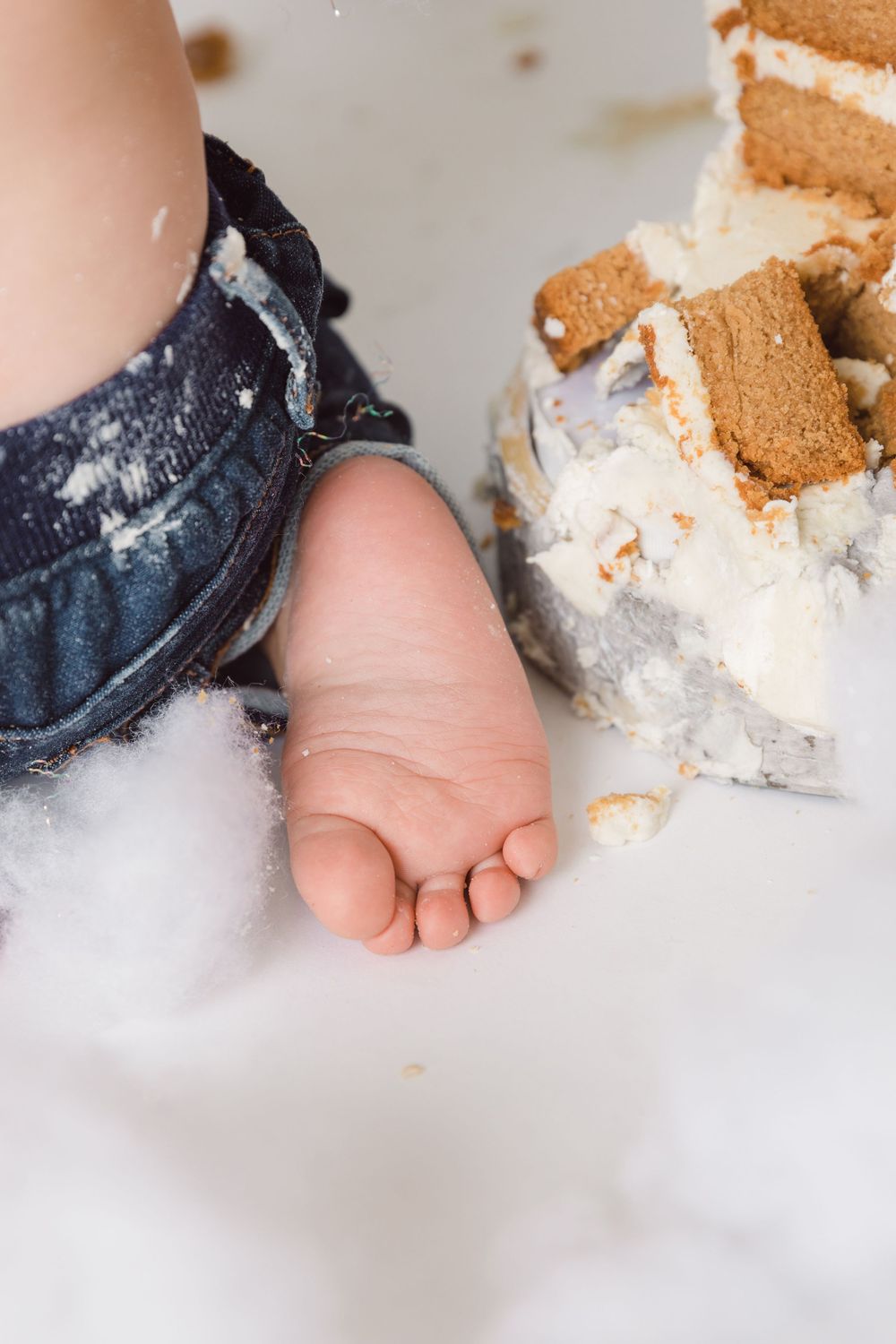 Baby's tiny foot next to crumbled cake and frosting on white surface during first birthday cake smash photo session.