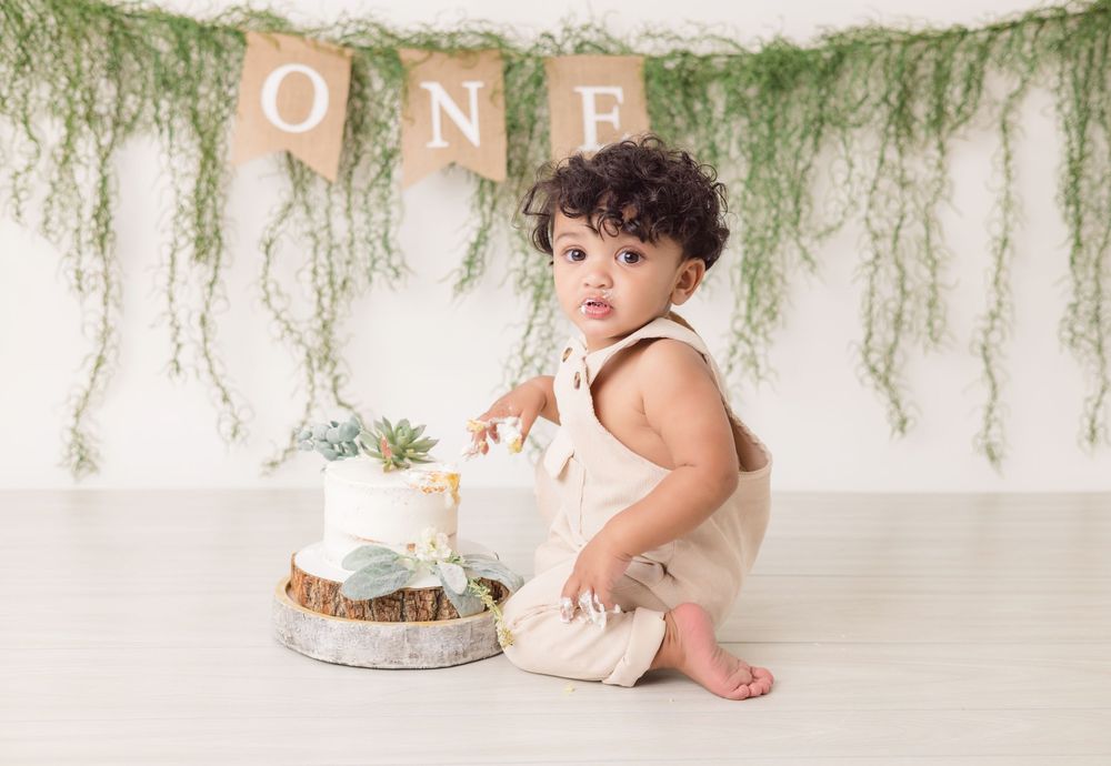A first birthday cake smash photo setup with a white cake, greenery backdrop, and 'ONE' banner decoration.