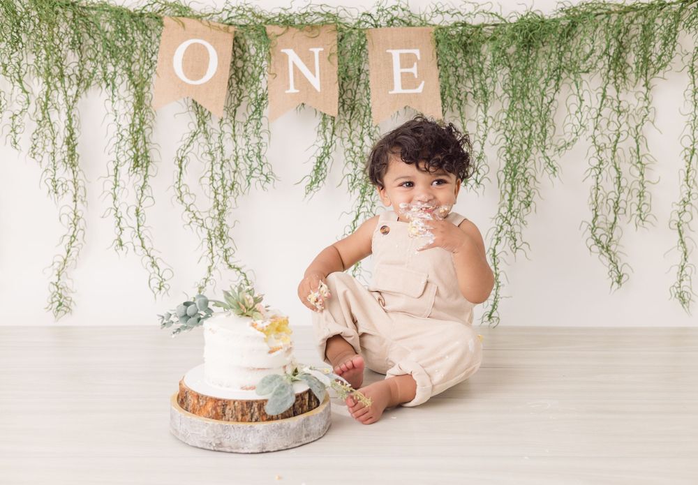 A baby celebrates their first birthday with a cake smash photo session under hanging greenery decorations.