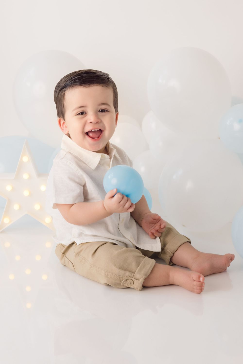 Adorable toddler in white shirt and khaki shorts sits on cloud backdrop with star light and blue balloon decor.