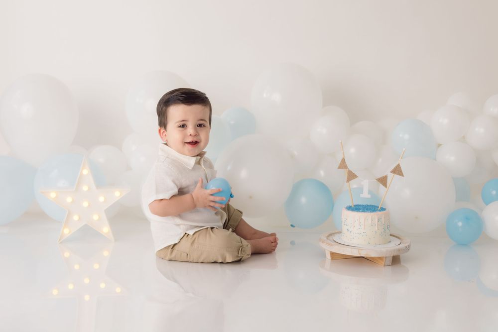 A baby sits on floor for cake smash photo session with blue balloons and twinkling star lights in background.