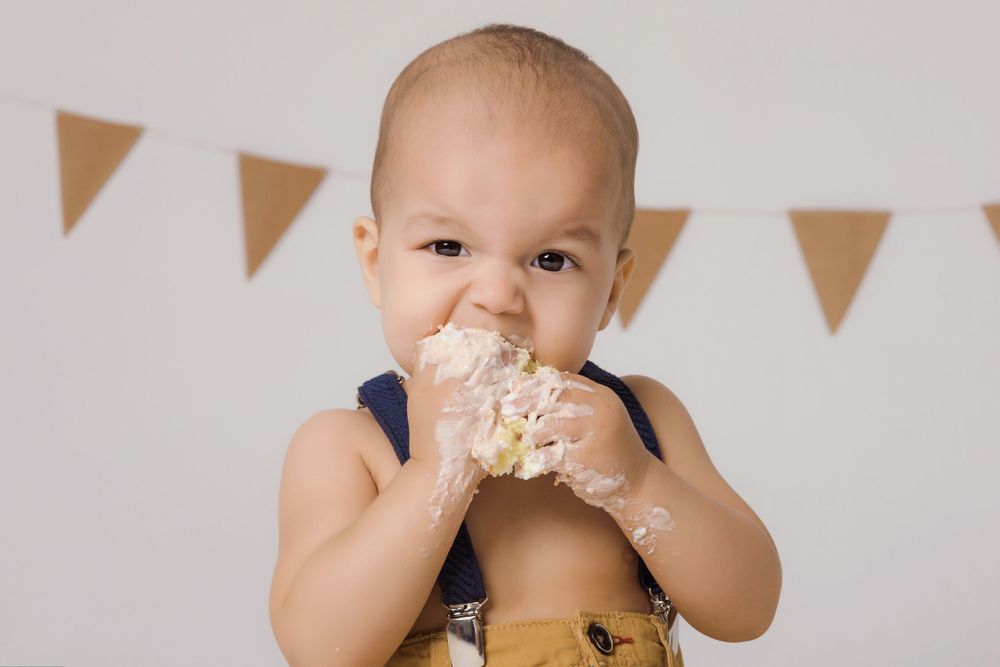 A baby enthusiastically enjoys eating cake during a first birthday celebration with colorful bunting decorations.