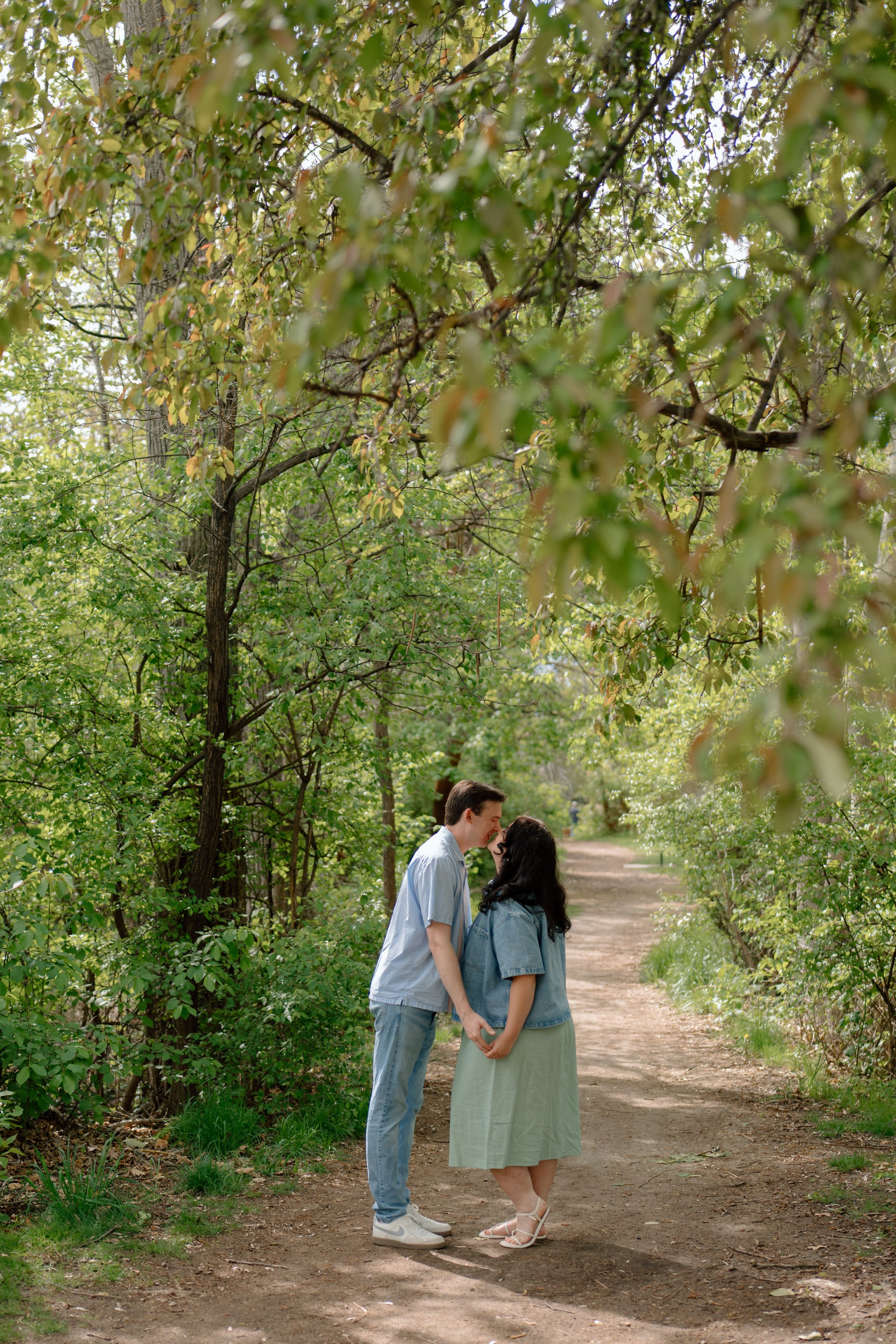 A romantic couple shares a tender moment on a tree-lined dirt path during a sunny autumn day.