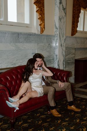 A couple shares an intimate moment on a vintage red velvet couch in a room with marble walls and ornate decor.