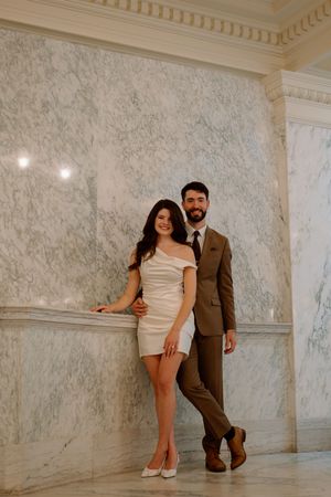 A couple poses elegantly against a marble wall, with her in a white dress and him in a brown suit.