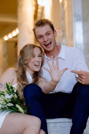 Newly engaged couple laughs joyfully while showing off ring and holding white floral bouquet in elegant setting.