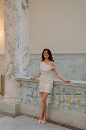 Person in white off-shoulder mini dress and heels poses elegantly against marble railing and columns in classic interior.