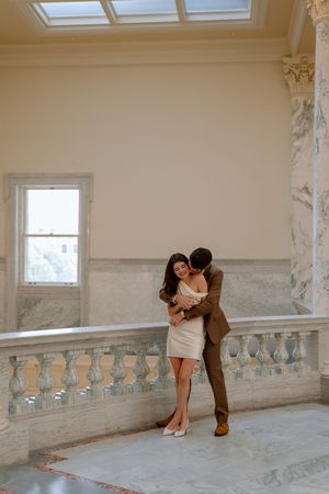 Romantic couple embraces in elegant marble hallway with skylight and vintage architectural details.