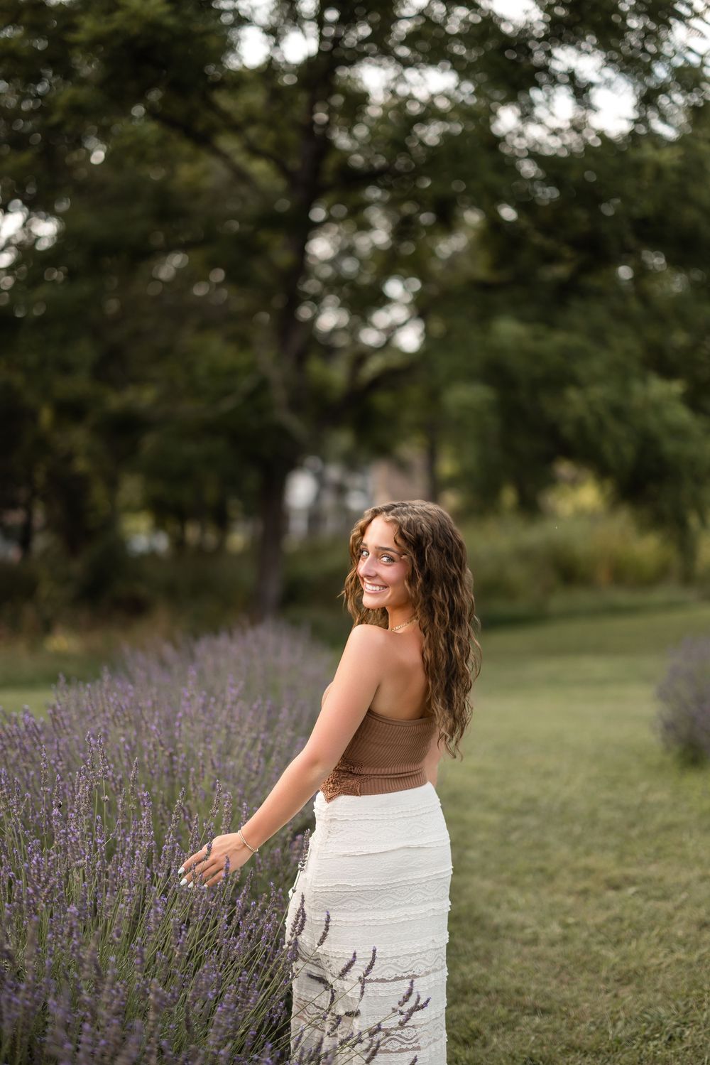 Someone in a brown top and white skirt stands beside blooming lavender in a garden setting at dusk.