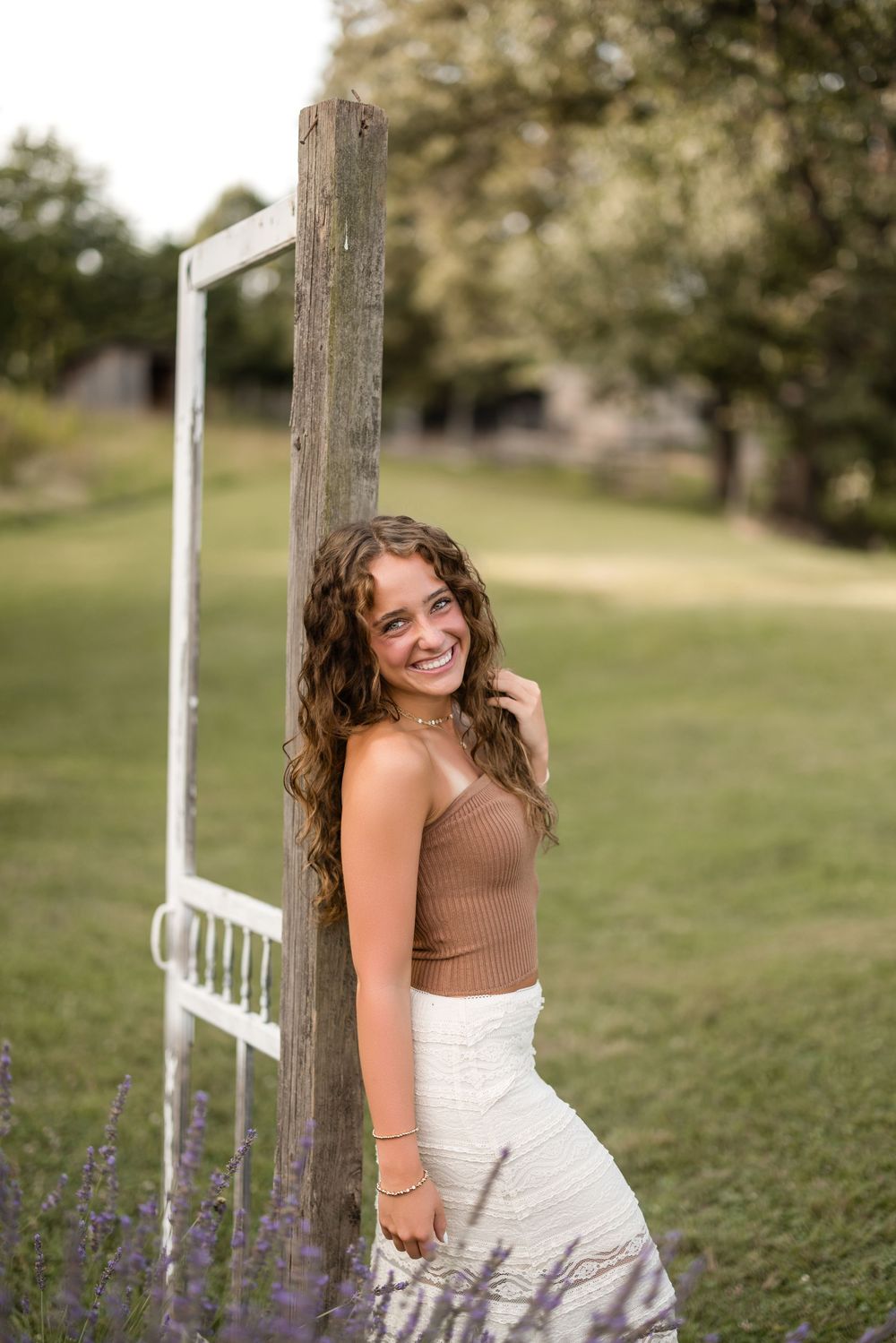 A young woman in a brown sweater and white skirt poses outdoors in a field with purple wildflowers and wooden fence posts.