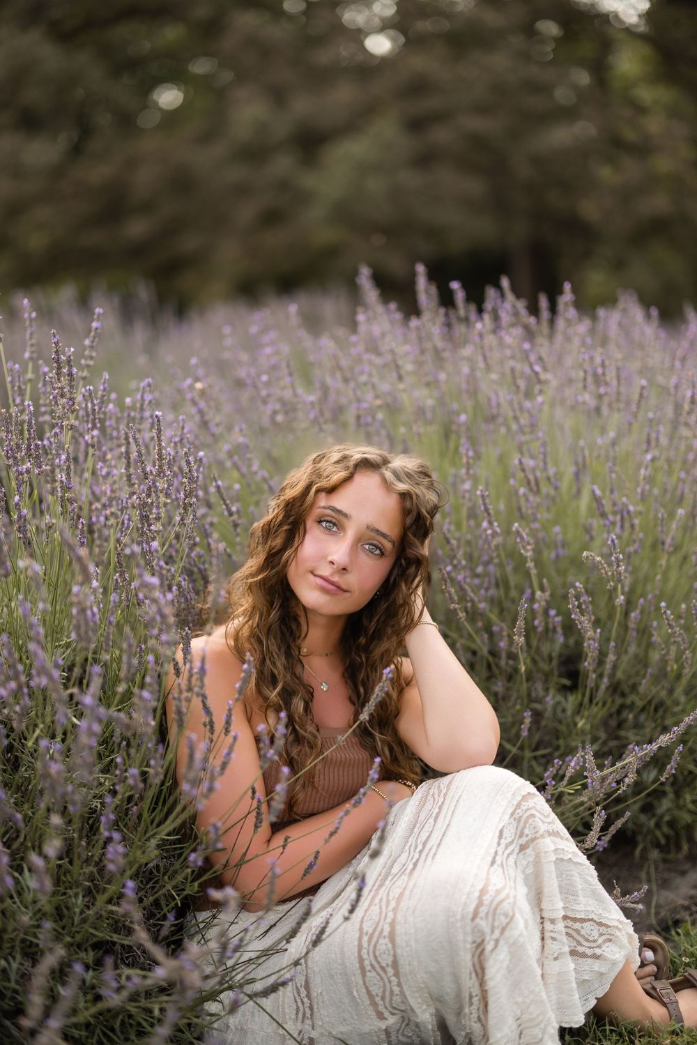 A woman in a white dress sits gracefully among blooming lavender flowers in a garden setting.