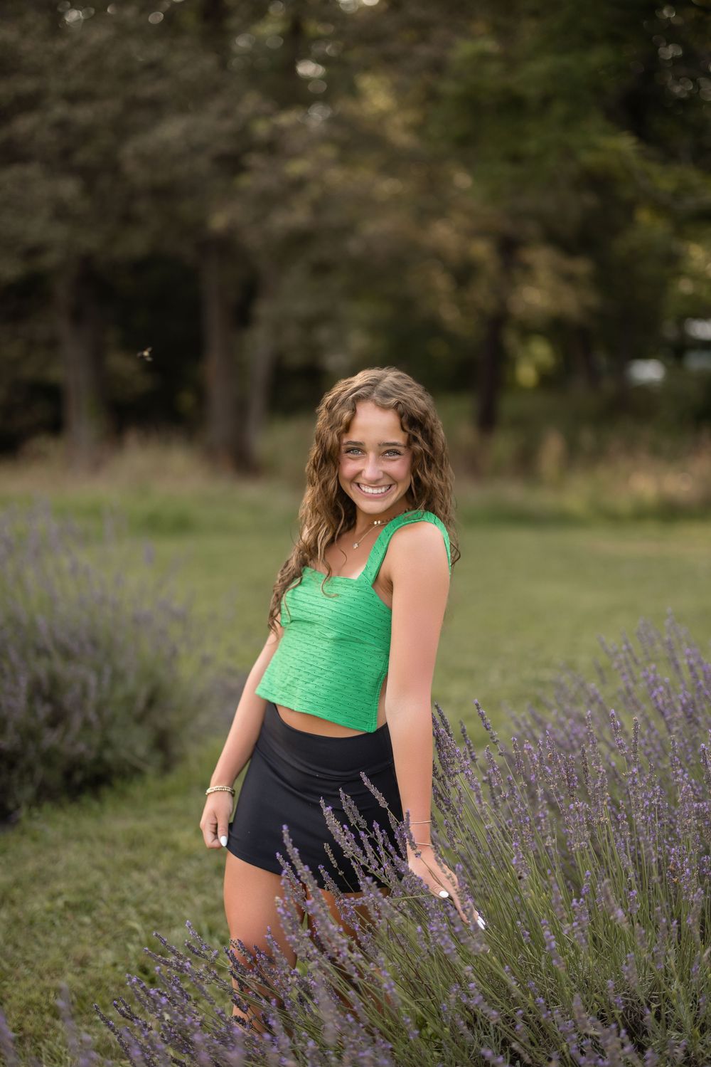 Person in bright green tank top and black shorts poses among blooming lavender flowers in a garden setting.