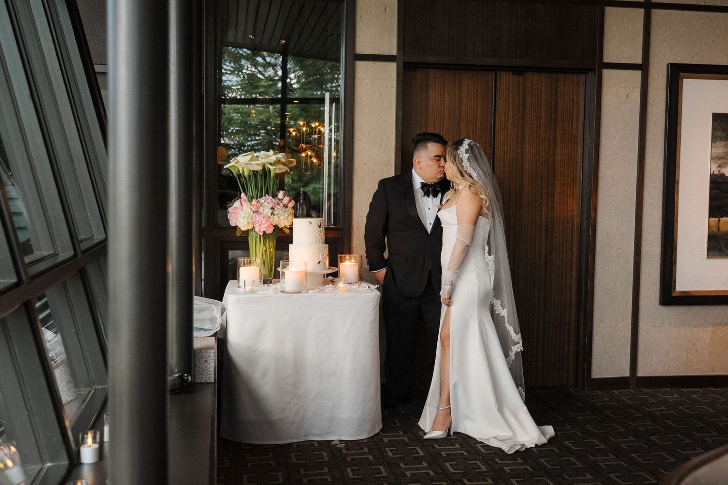 A couple in wedding attire stands near an elegant table with floral arrangements and candles by a window.
