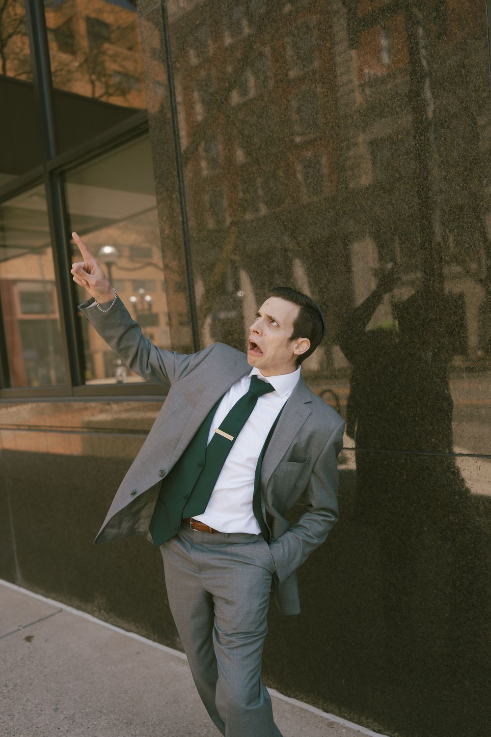 A person in a grey suit strikes dramatic poses against a dark brick wall backdrop.