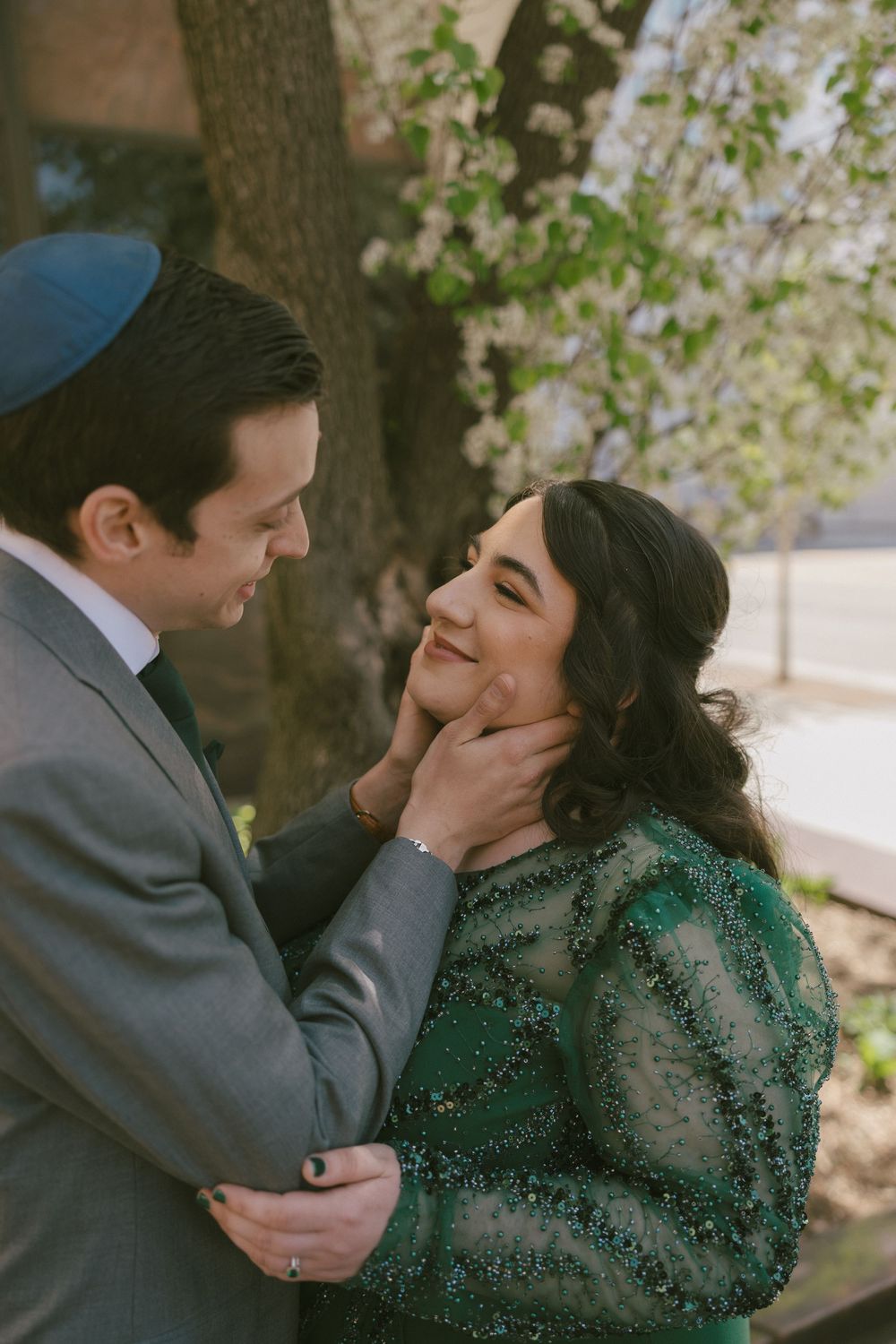 A romantic couple embracing outdoors under blooming cherry blossoms wearing formal attire.