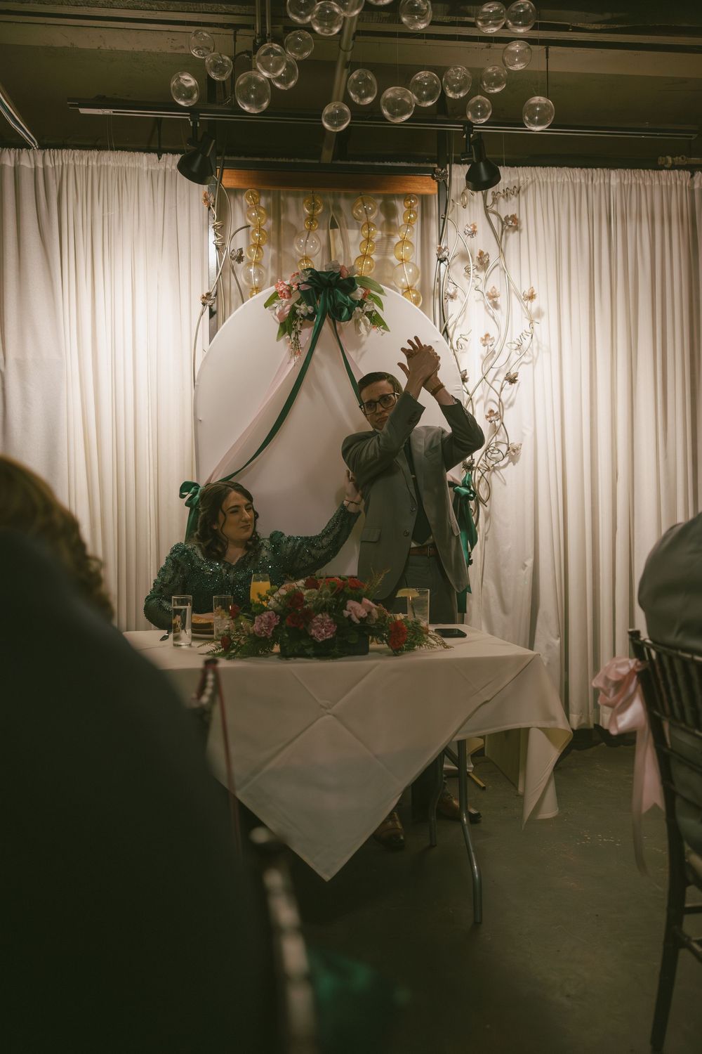 Wedding reception venue with white drapes, hanging lights and guests celebrating at decorated tables.