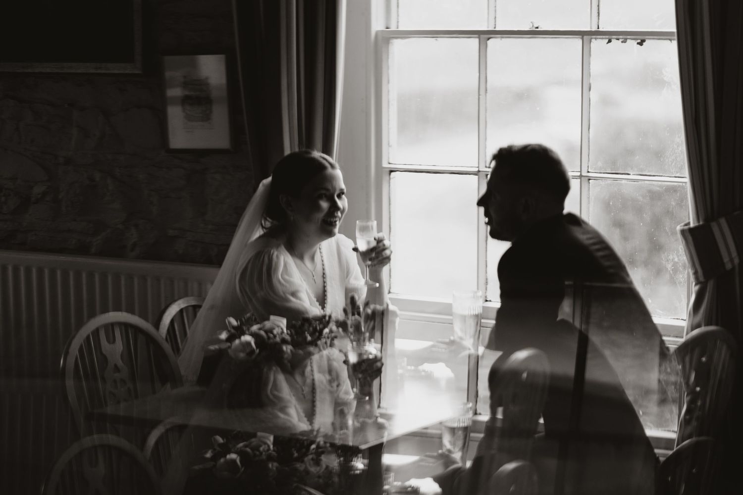 Two people sit in silhouette by a window in a moody black and white photograph during an intimate conversation.