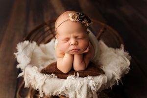 Sleeping newborn in brown headband rests peacefully on white blanket in wooden bowl during professional photoshoot.