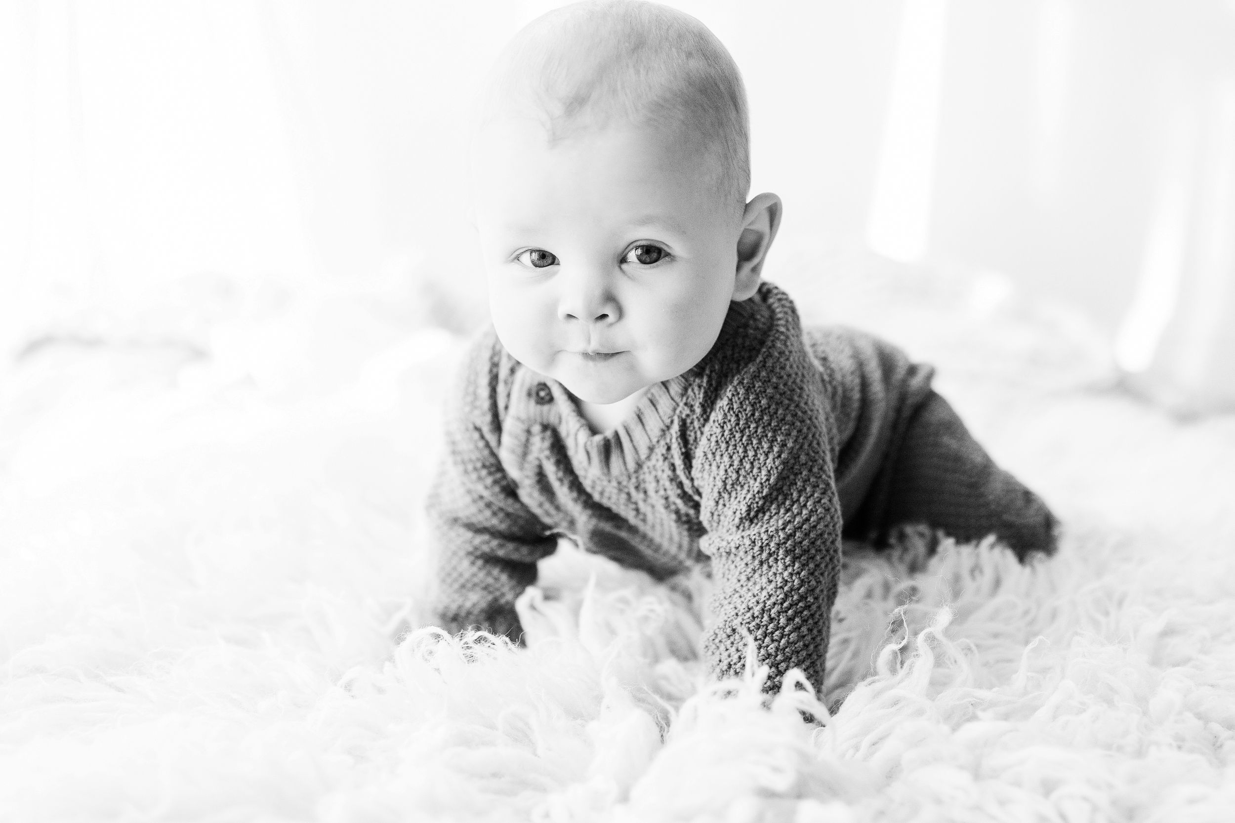 Black and white portrait of a baby lying on a fluffy white rug wearing a knit sweater.