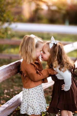 Two children in brown and white dresses play together on a wooden fence in an autumn setting.