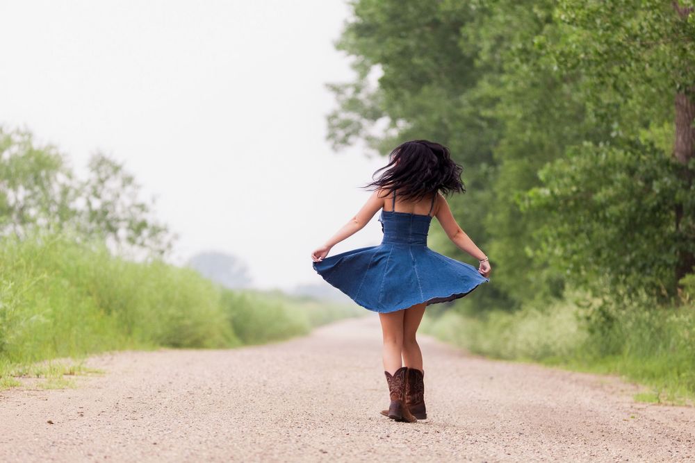 Person in a blue dress walks away on a dirt path through lush green countryside on a sunny day.