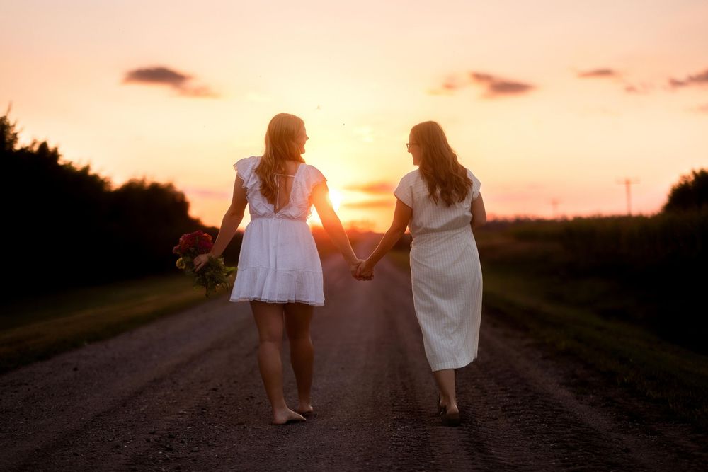 Two people in white dresses walk down a dirt road at sunset while holding hands.