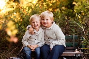 Two young children in light colored shirts sit together outdoors during golden hour lighting.