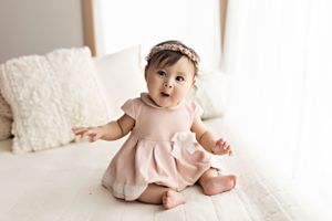 Baby in pink dress sitting on white bedding with surprised expression.