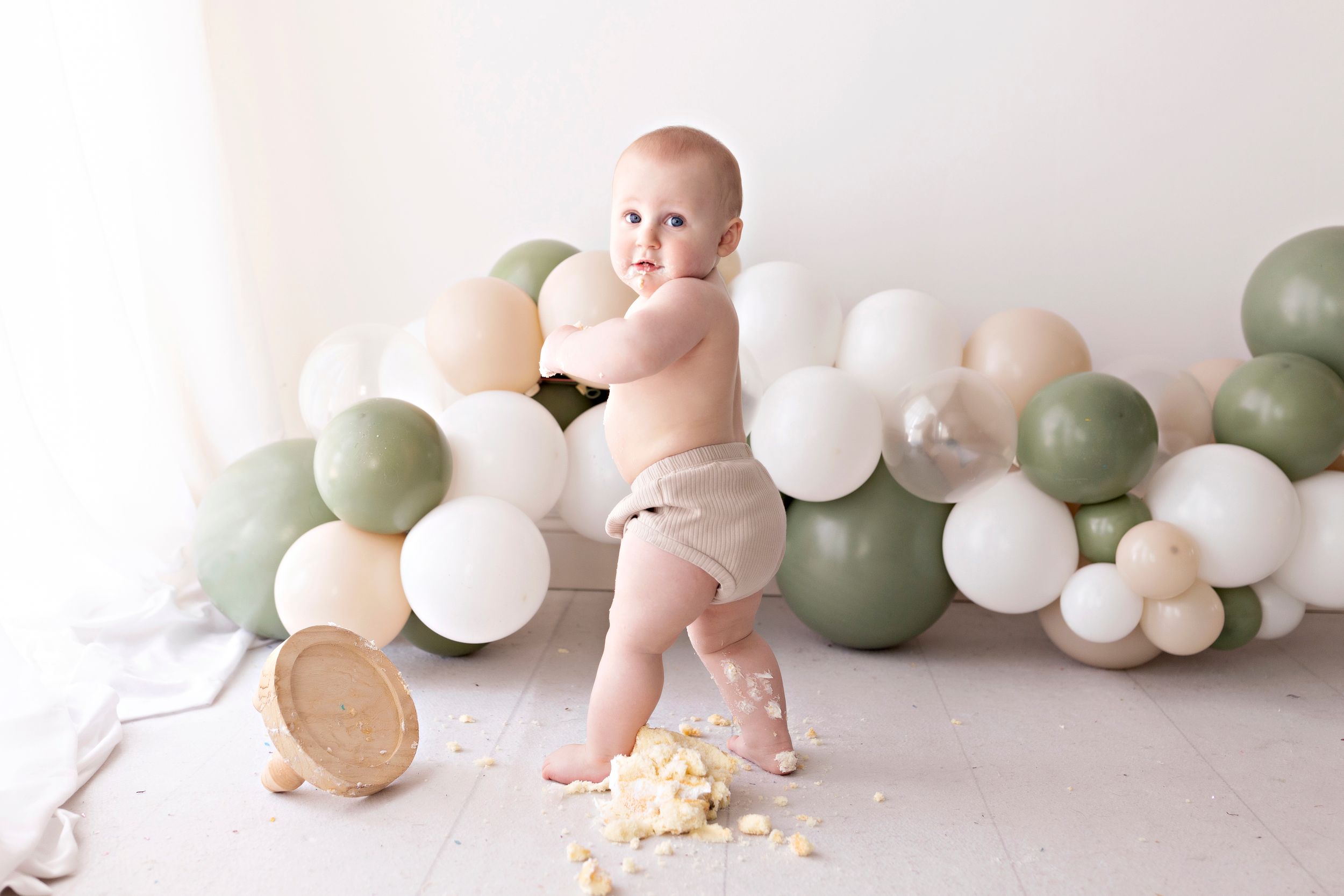 Baby in diaper plays with cake amid green and white balloon decorations.