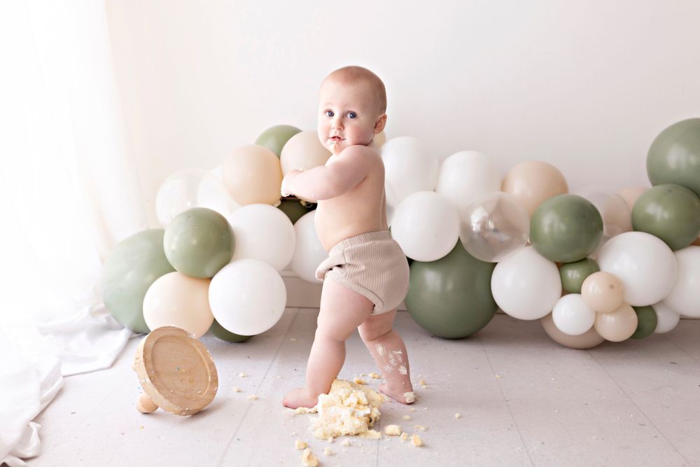 Baby in diaper plays with cake amid green and white balloon decorations.