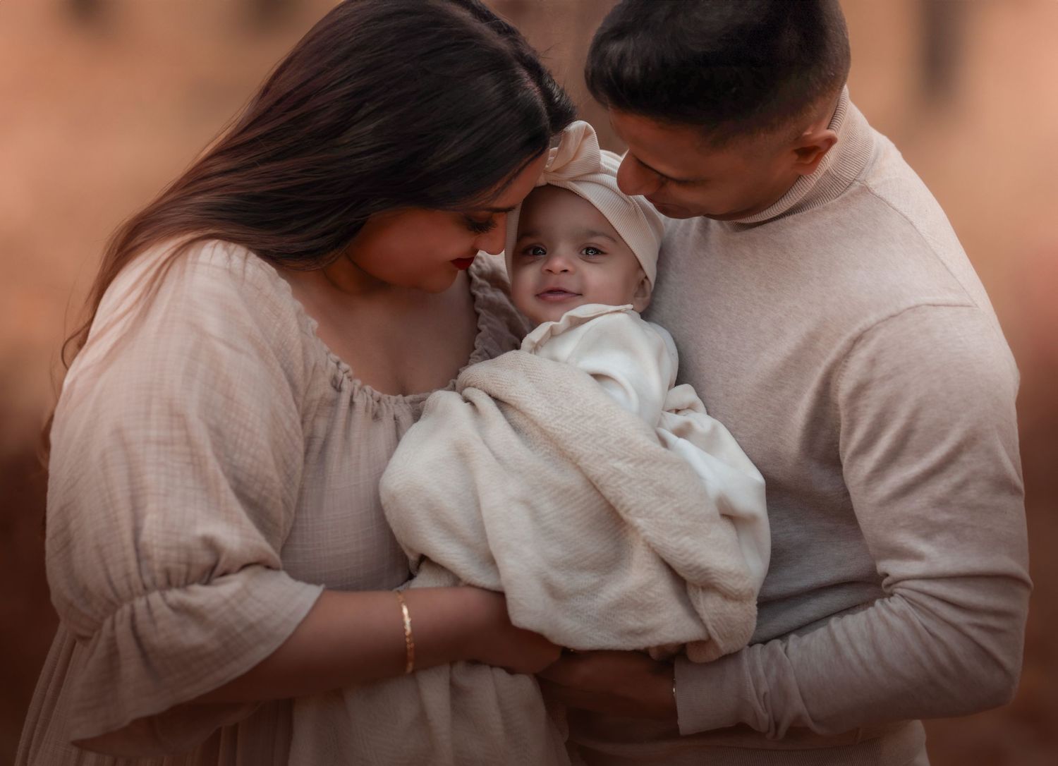 A couple tenderly holds their newborn baby wrapped in white, sharing an intimate family moment outdoors.