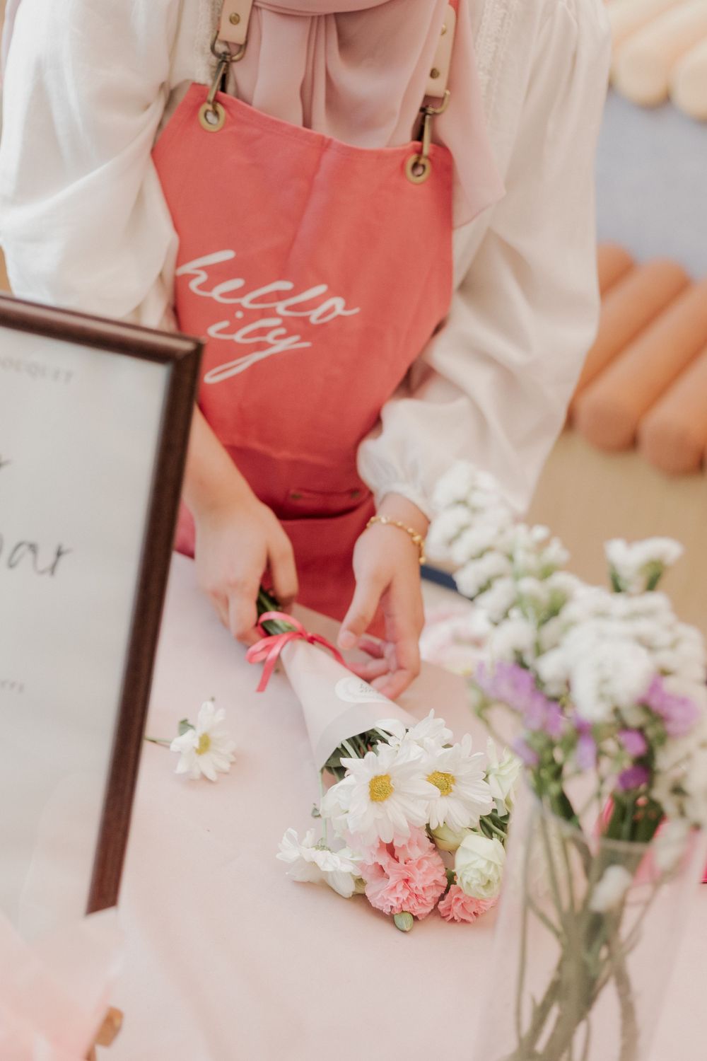 Close-up of hands arranging flowers with pink apron and framed signage visible.