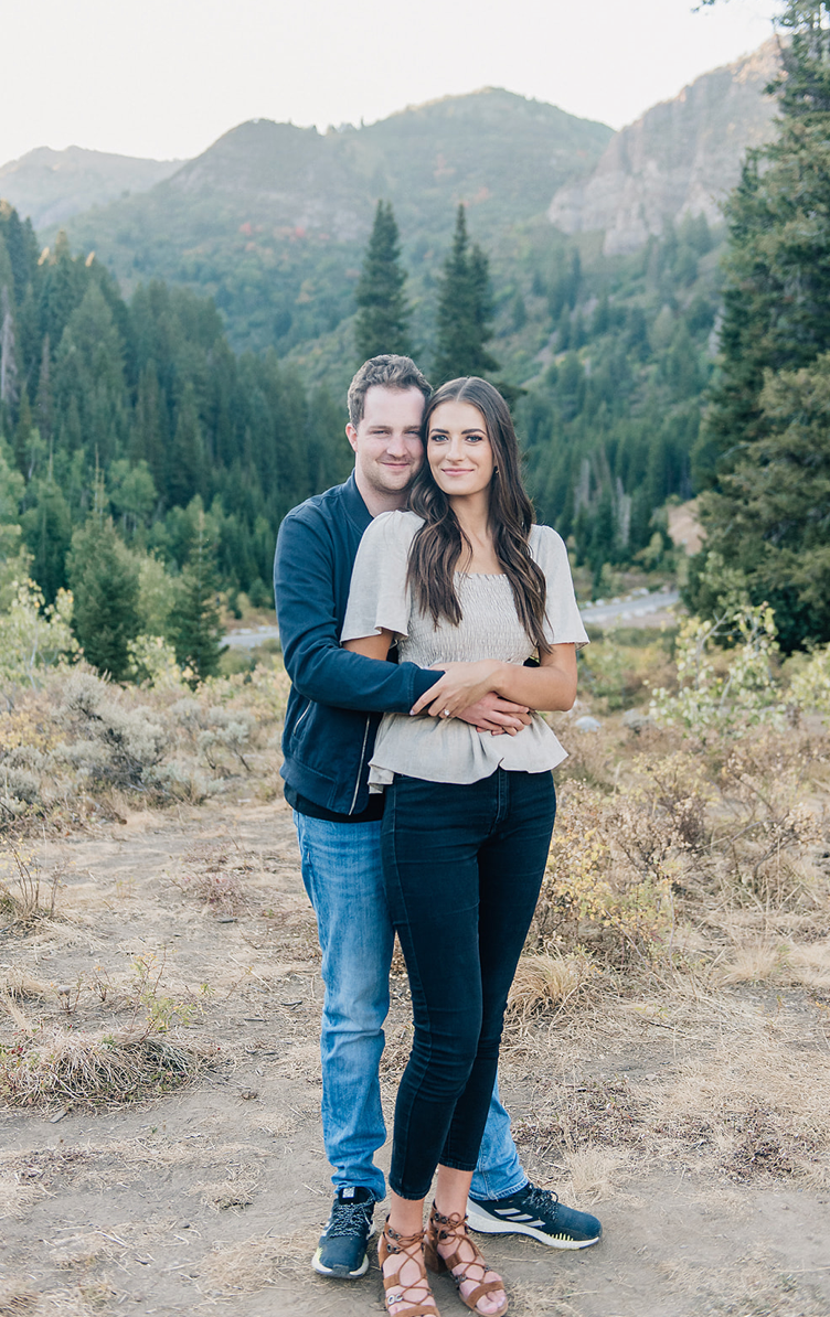 Couple poses for engagement photo with mountain landscape and pine trees in background.