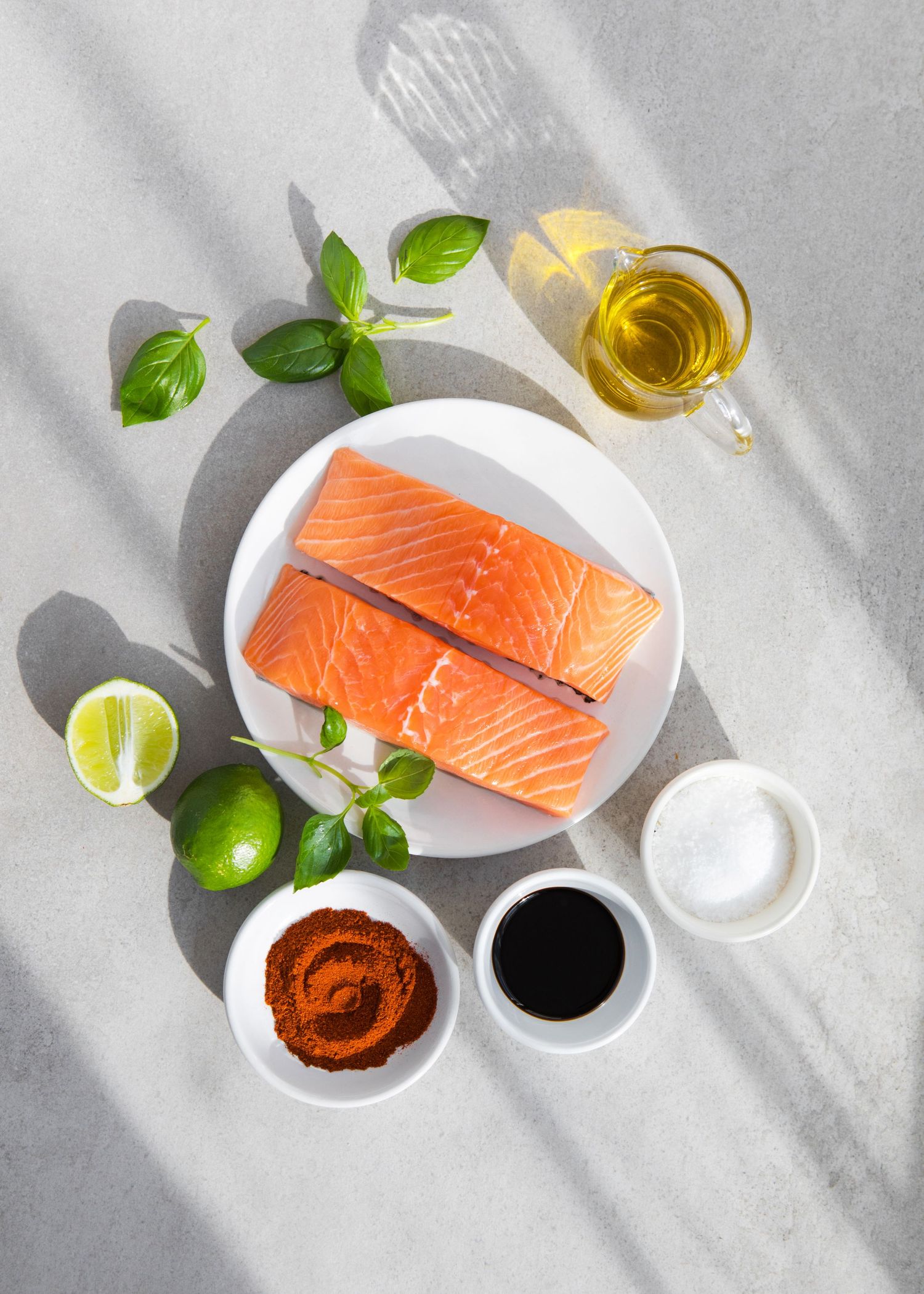Two raw salmon fillets on a white plate surrounded by fresh basil, lime, spices, soy sauce, oil, and salt.