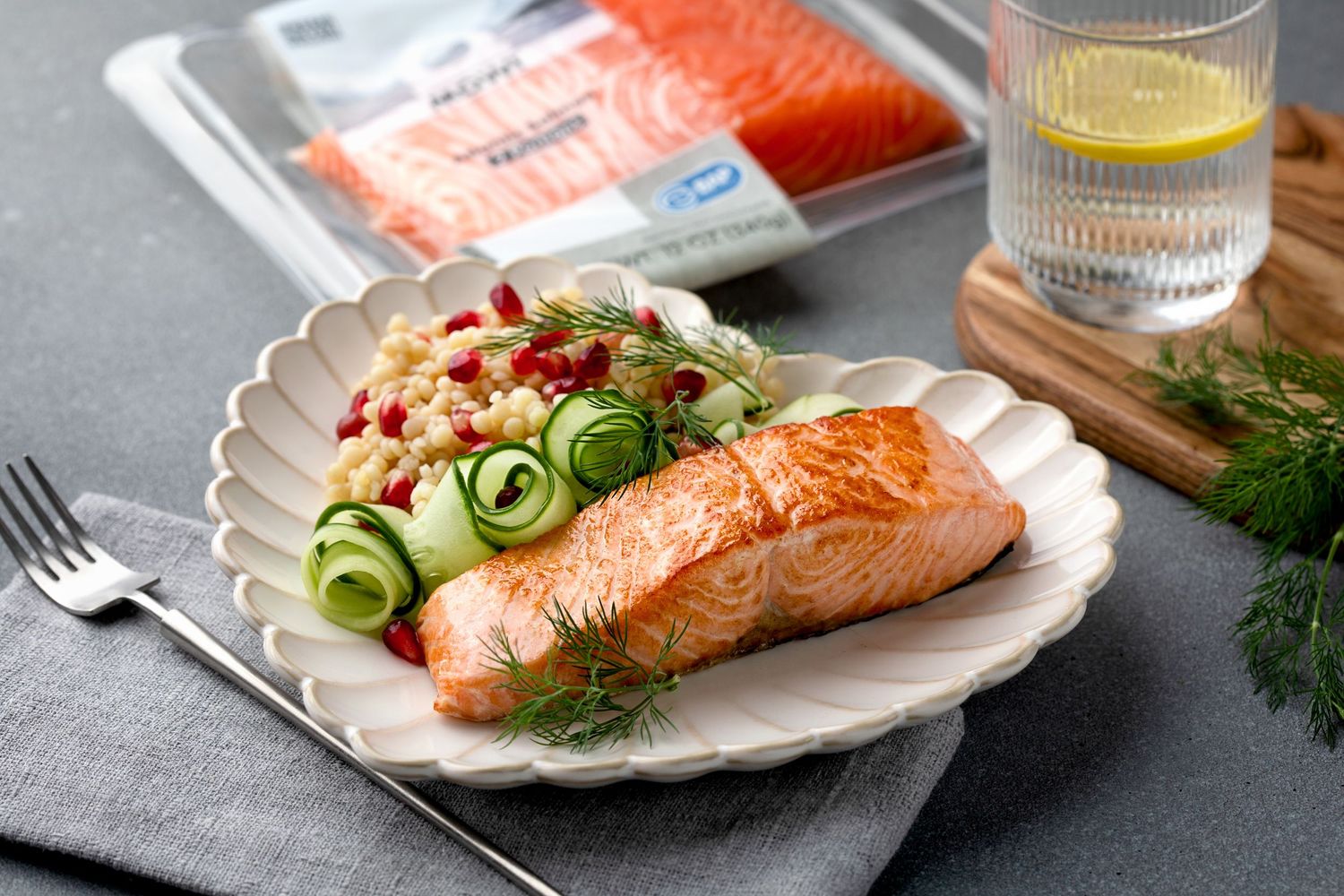 Grilled salmon fillet served with cucumber salad, pomegranate, dill, and couscous on a white scalloped plate.