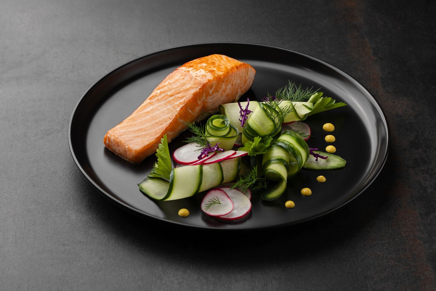 Grilled salmon fillet on a black plate with fresh cucumber, radish slices, dill, and yellow peas salad.