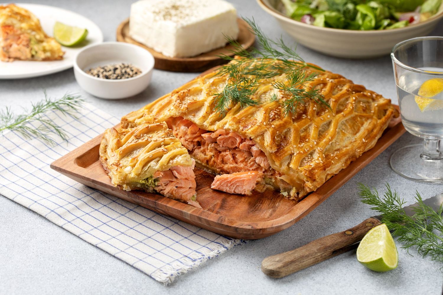 Golden puff pastry salmon en croute on wooden board with dill, cream cheese, and lemon wedges on light background.