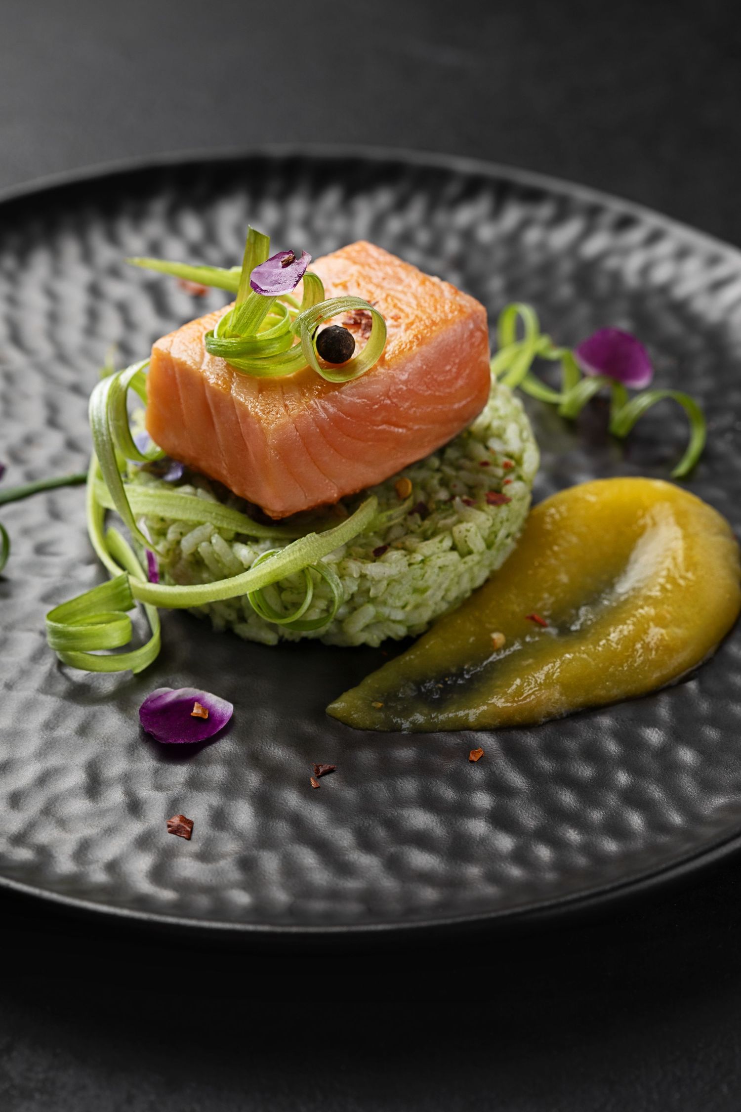 Elegant salmon fillet on green herb rice with pickled pear, microgreens, and edible flowers on a dark plate.