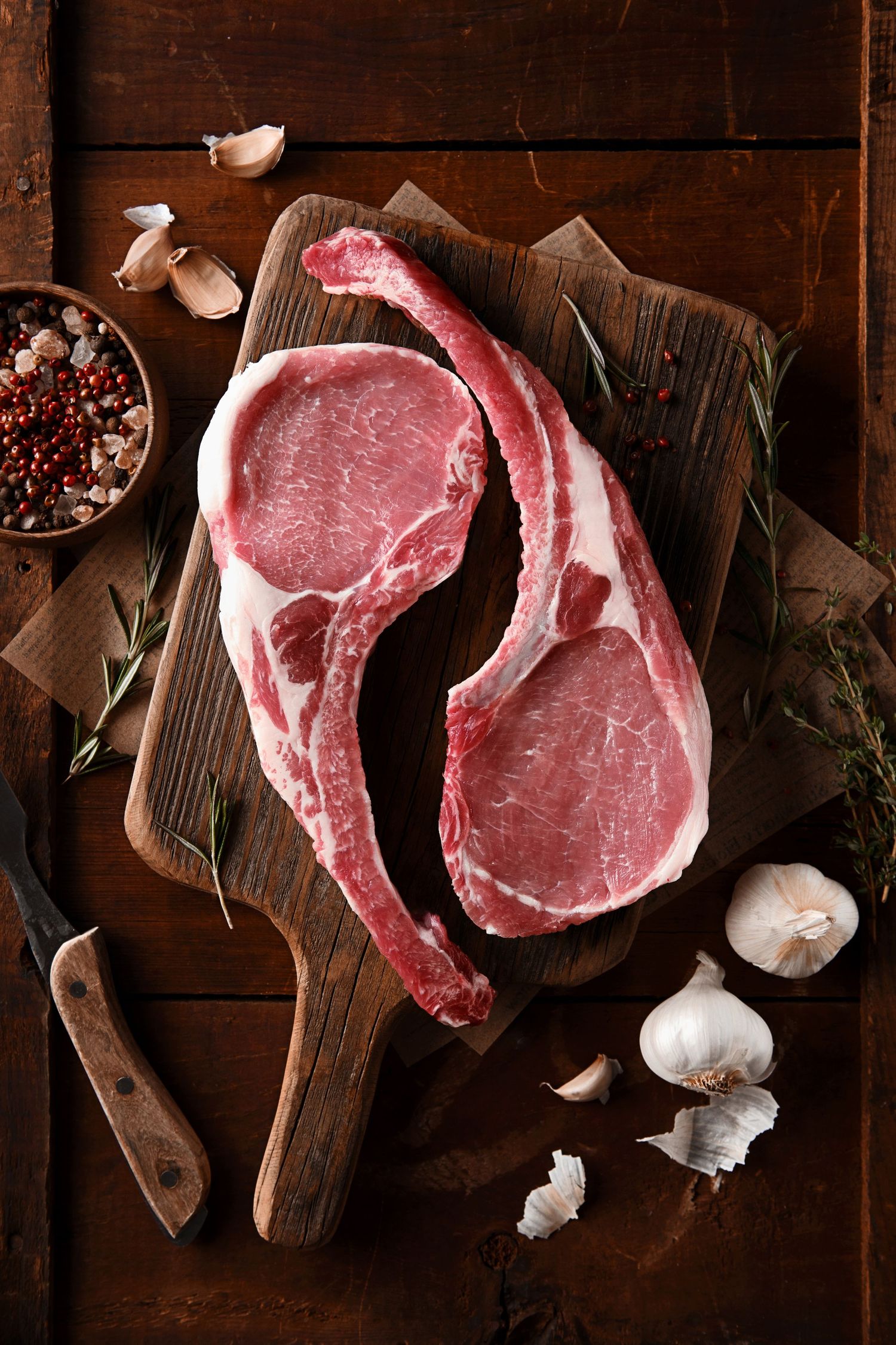 Raw bone-in pork chops arranged on rustic wooden cutting boards with garlic cloves and spices in dark lighting.