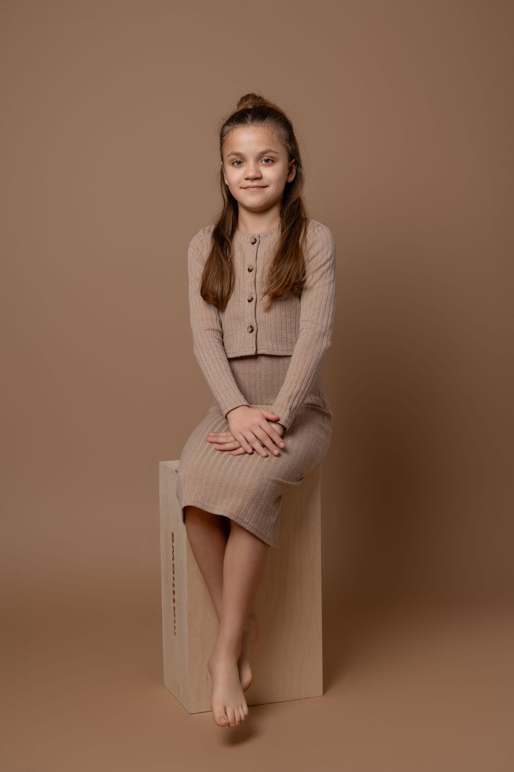 Portrait in neutral tones featuring ribbed beige dress and cardigan while seated on wooden box against brown backdrop.